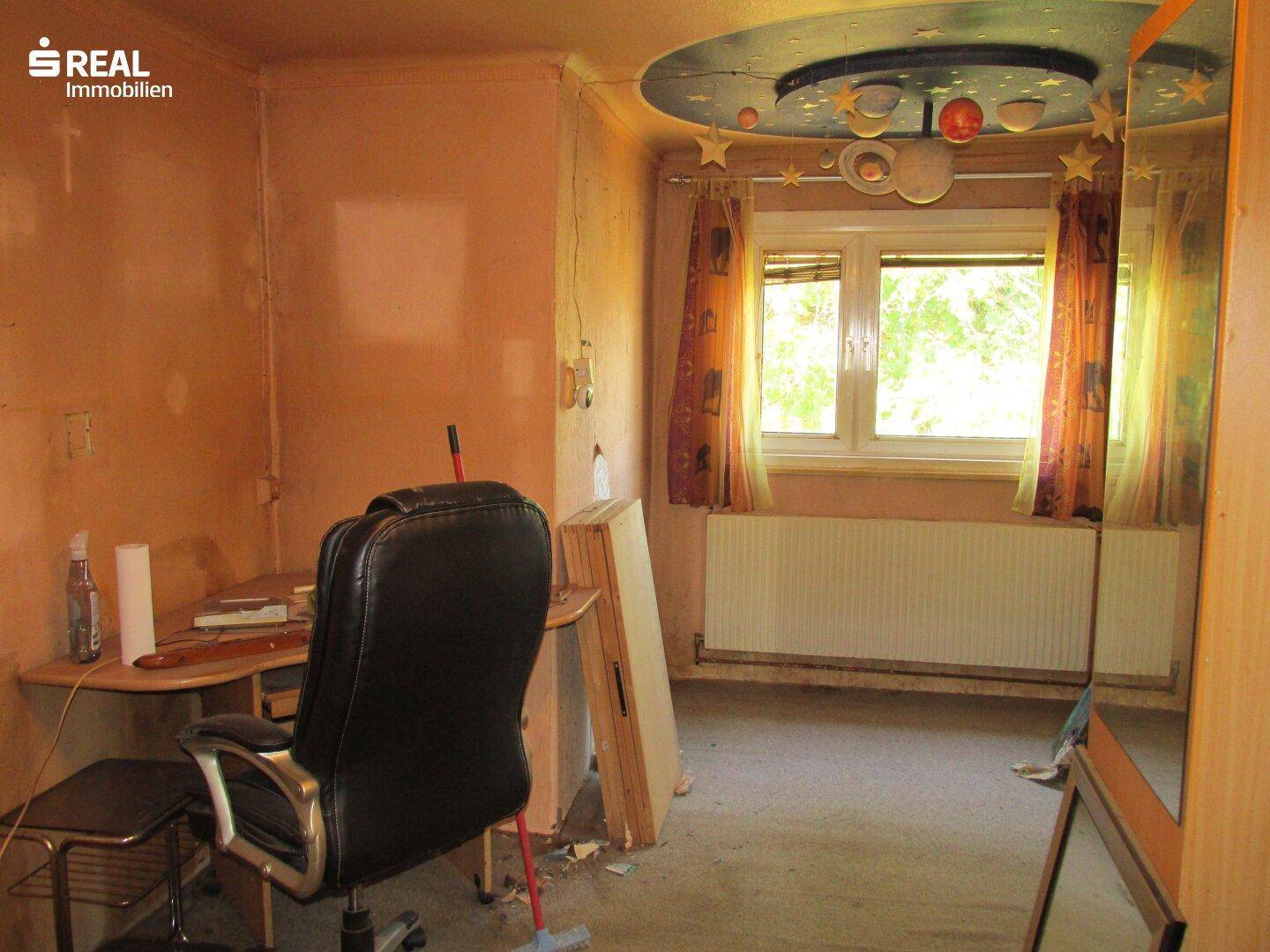 Zimmer mit Schreibtisch, Bürostuhl und einem Fenster, das viel natürliches Licht hereinlässt und eine grüne Aussicht bietet.
