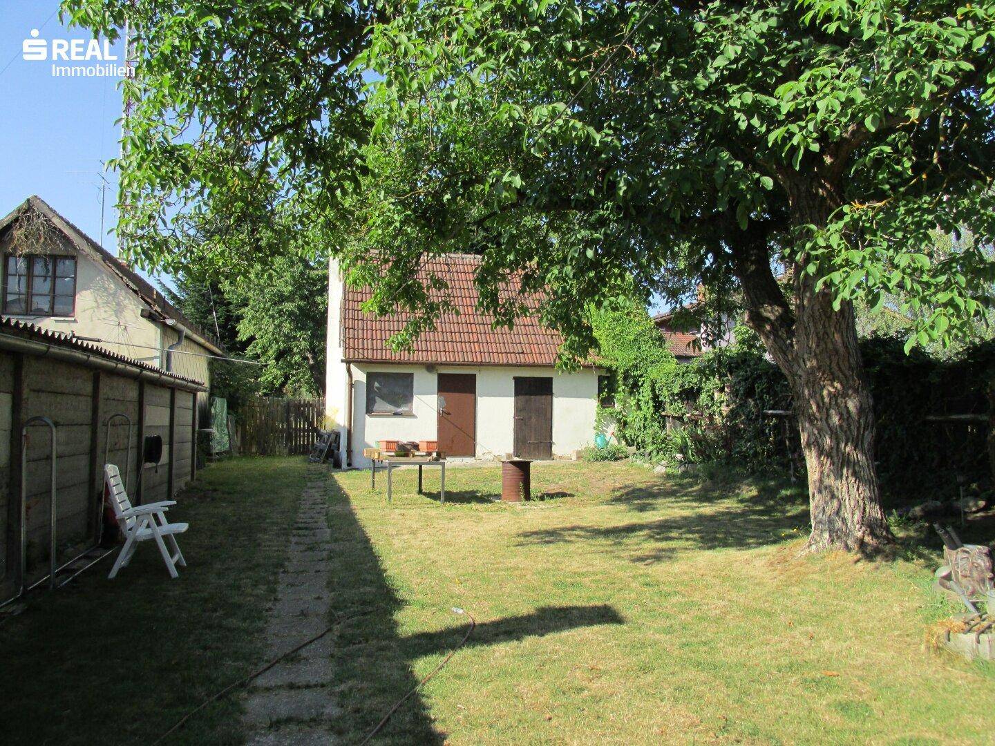 Großer Garten mit Rasen, altem Baum und einem kleinen Nebengebäude im Hintergrund.