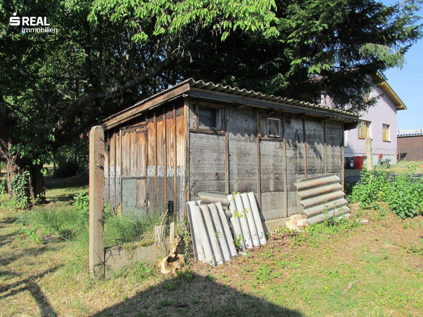 Rustikaler Holzschuppen im Garten mit Wellblechdach und umgebender Vegetation.