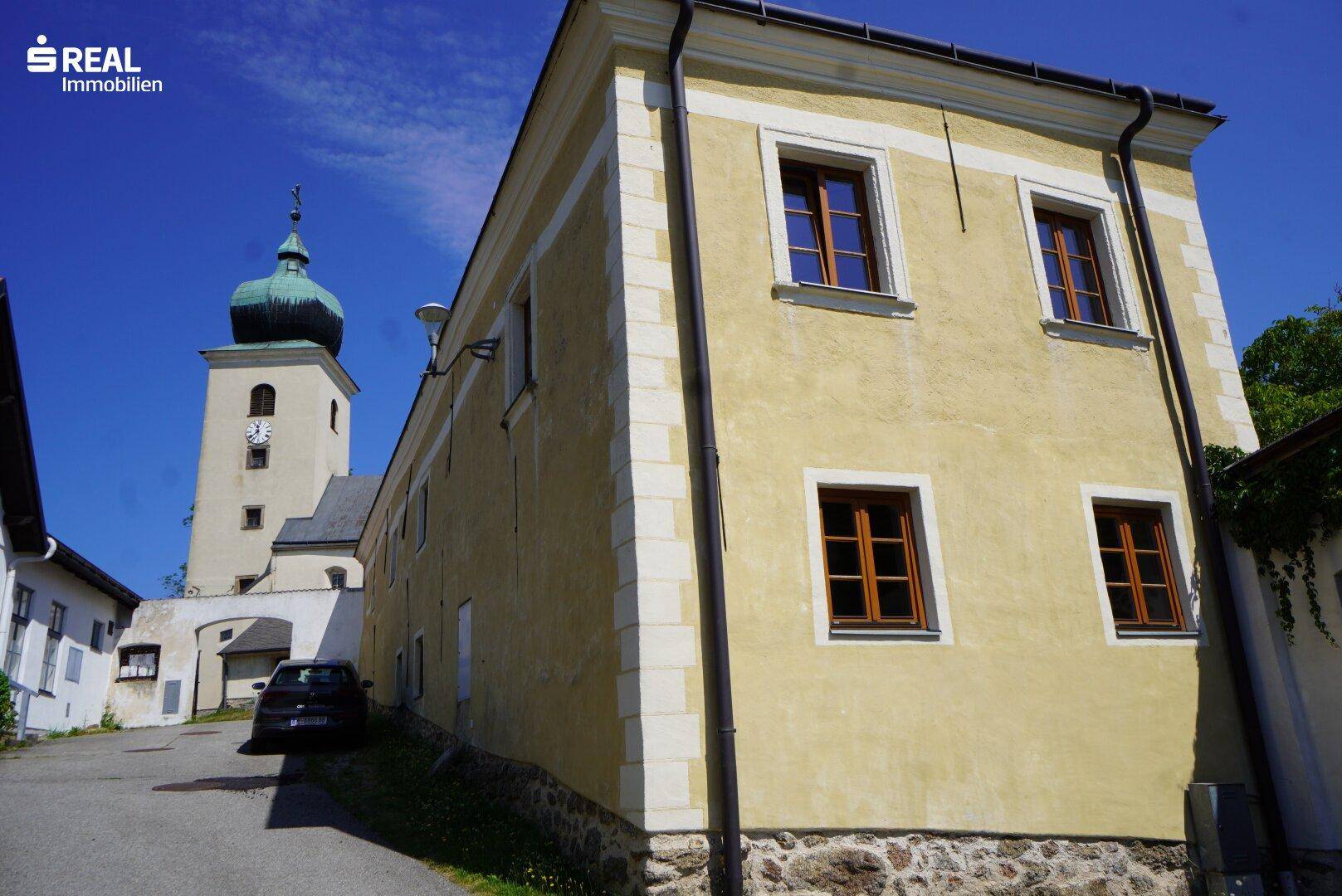 Seitenansicht des historischen Gebäudes mit gelber Fassade und einem Kirchturm im Hintergrund.