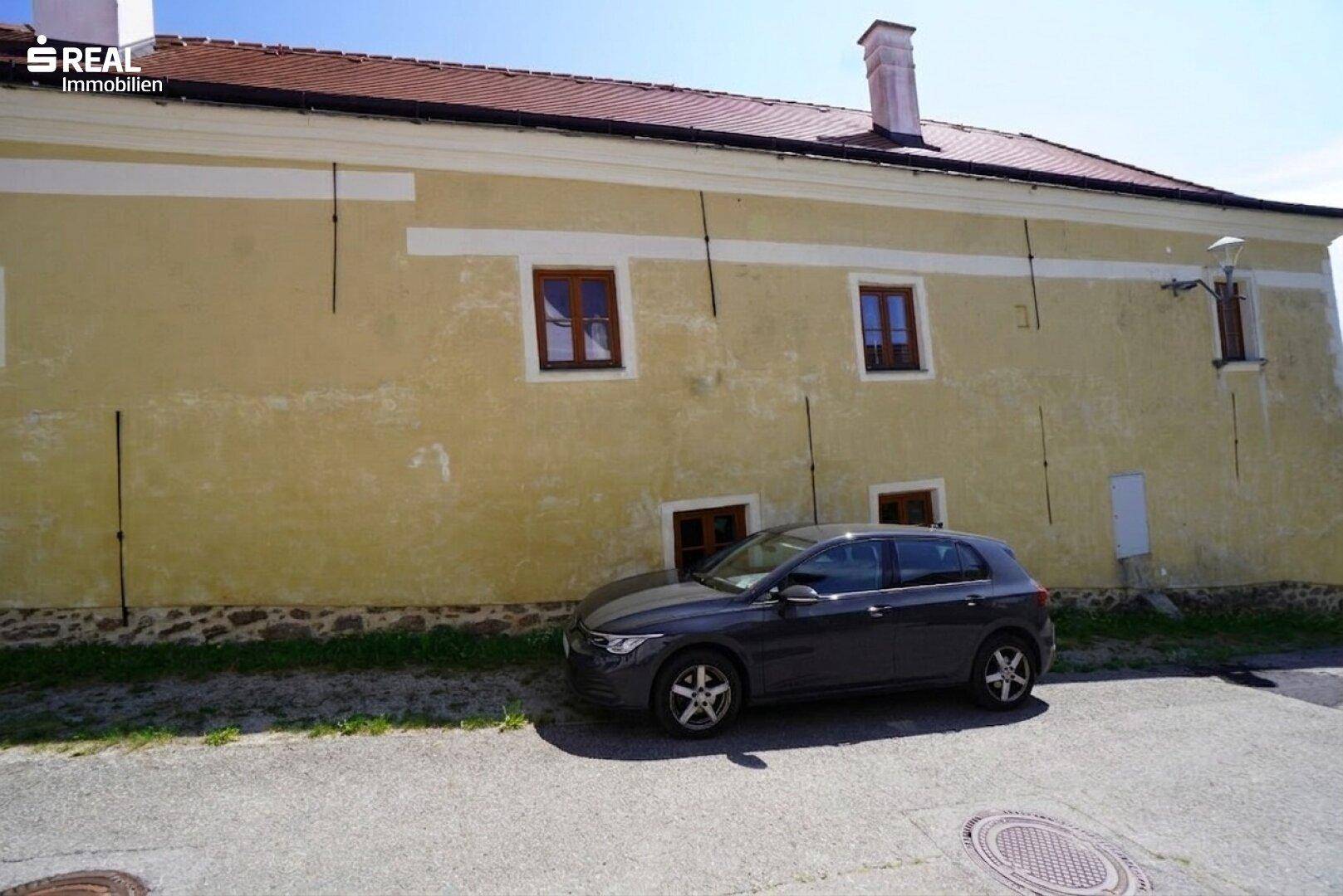 Ansicht der Gebäudeseite mit gelber Fassade und einem geparkten Auto auf dem Vorplatz.