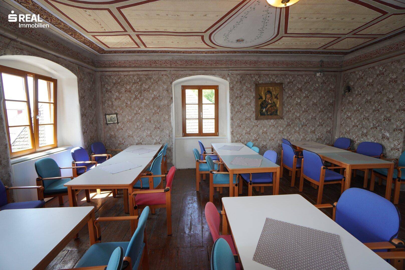 Traditioneller Speisesaal mit Holzboden, bemalter Decke und mehreren Fenstern.