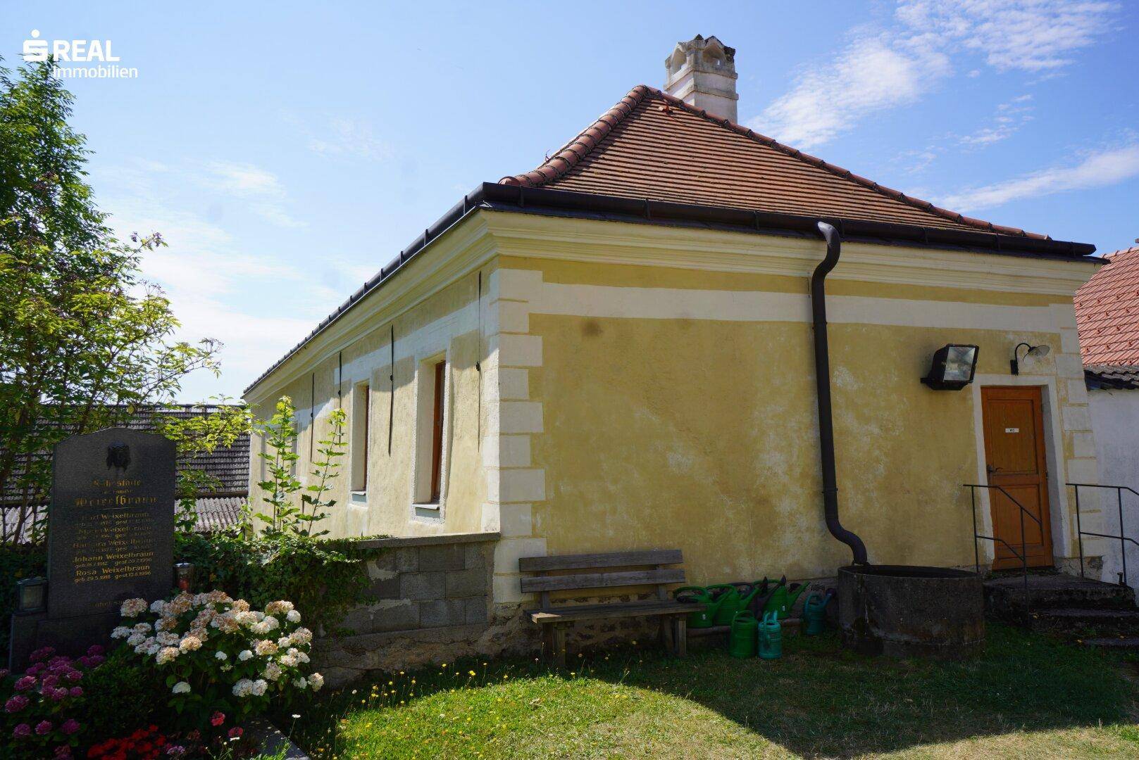 Kleineres Gebäude mit gelber Fassade, einer Holzbank und einem Gedenkstein im Vorgarten.