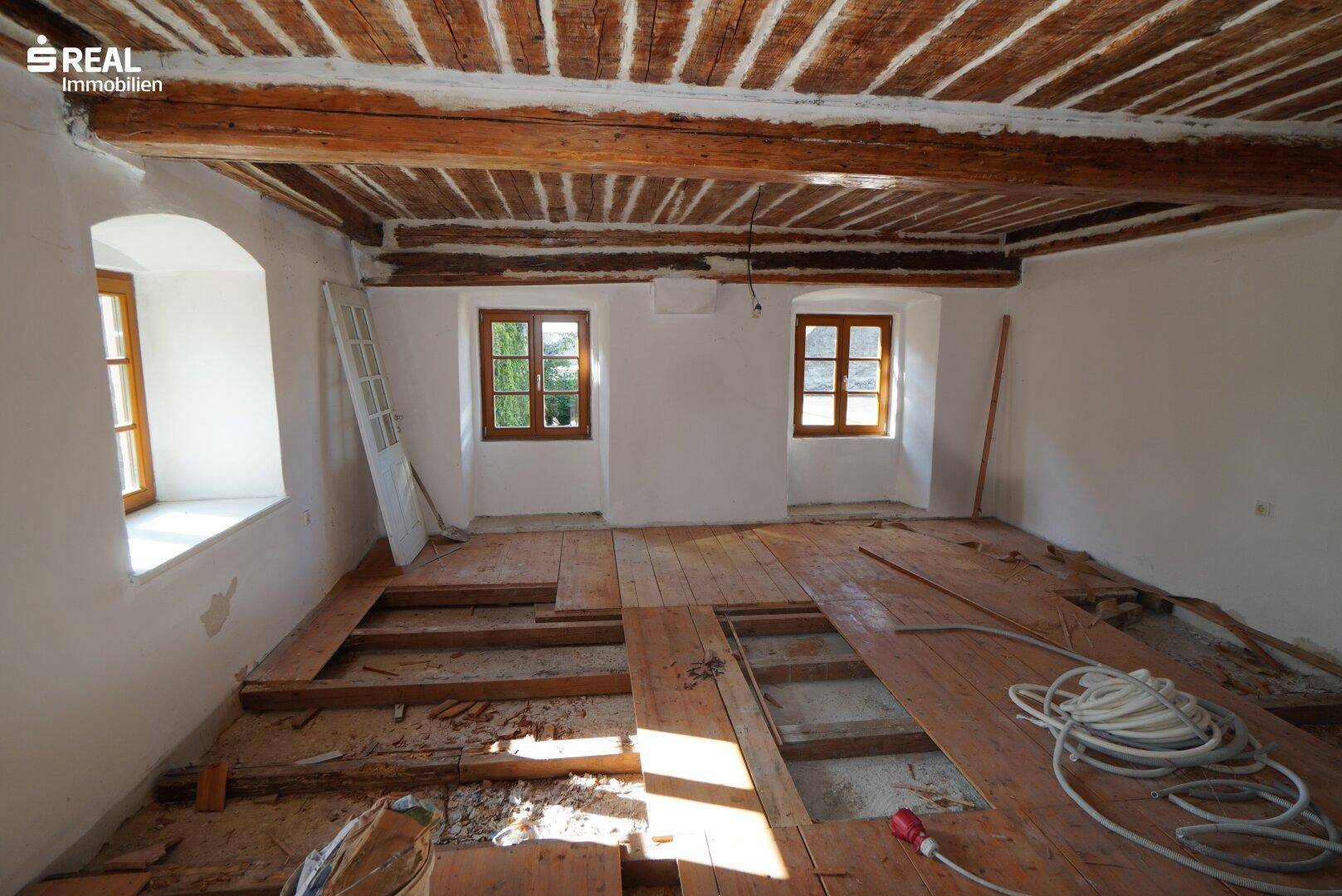Renovierungsbedürftiger Raum mit freiliegenden Holzbalken an der Decke und Dielenboden.