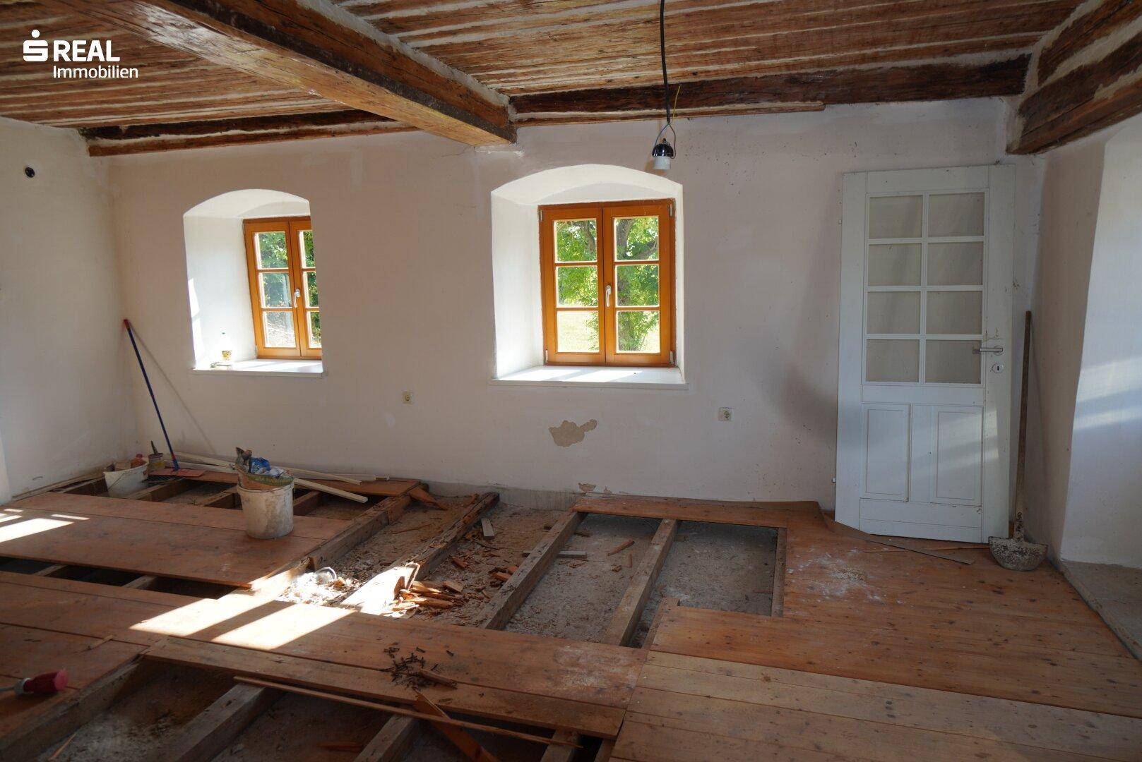 Renovierungsbedürftiger Raum mit Holzbalkendecke, Dielenboden und zwei Fenstern.