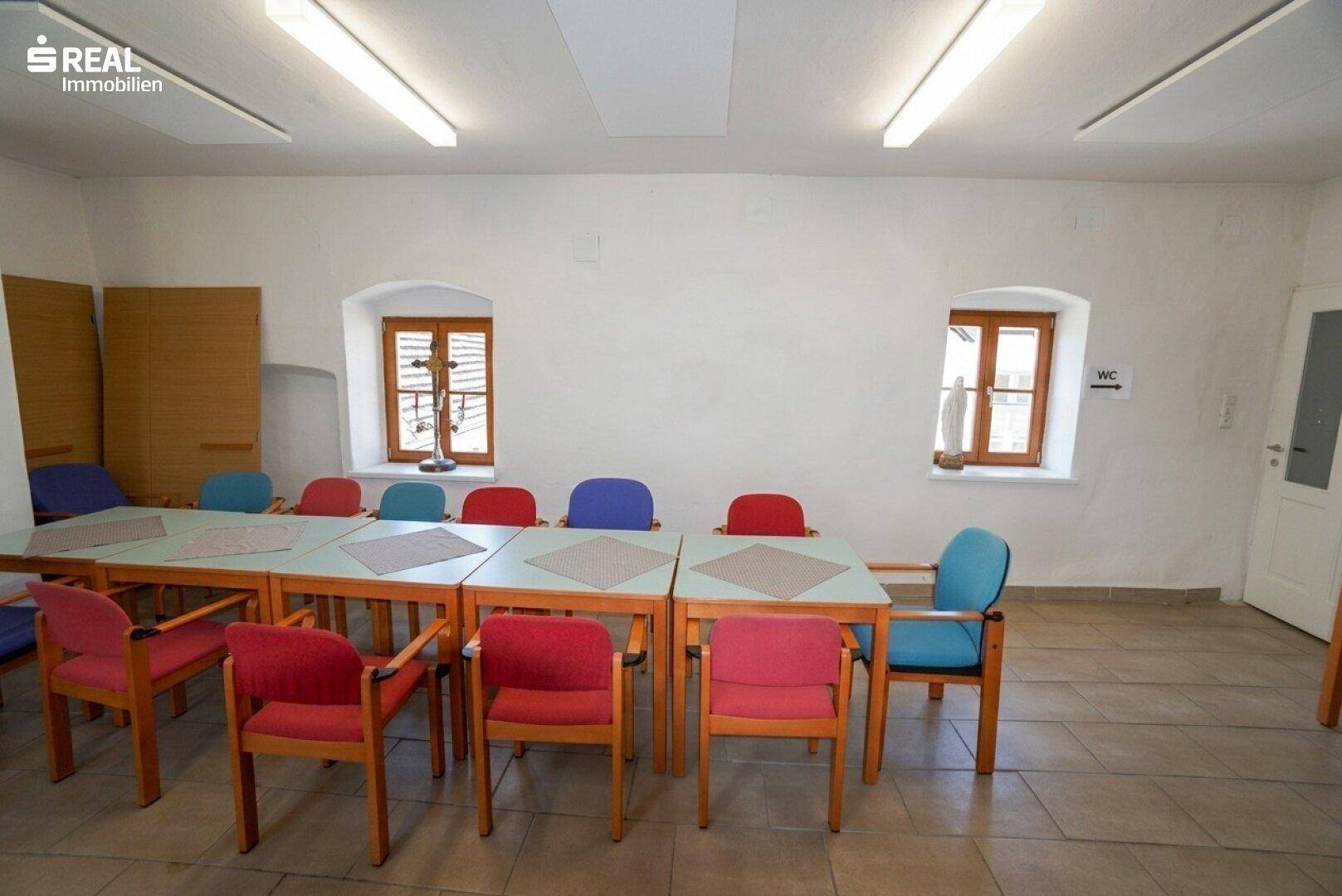 Einfacher Essbereich mit mehreren Tischen und Stühlen, beleuchtet durch kleine Fenster.