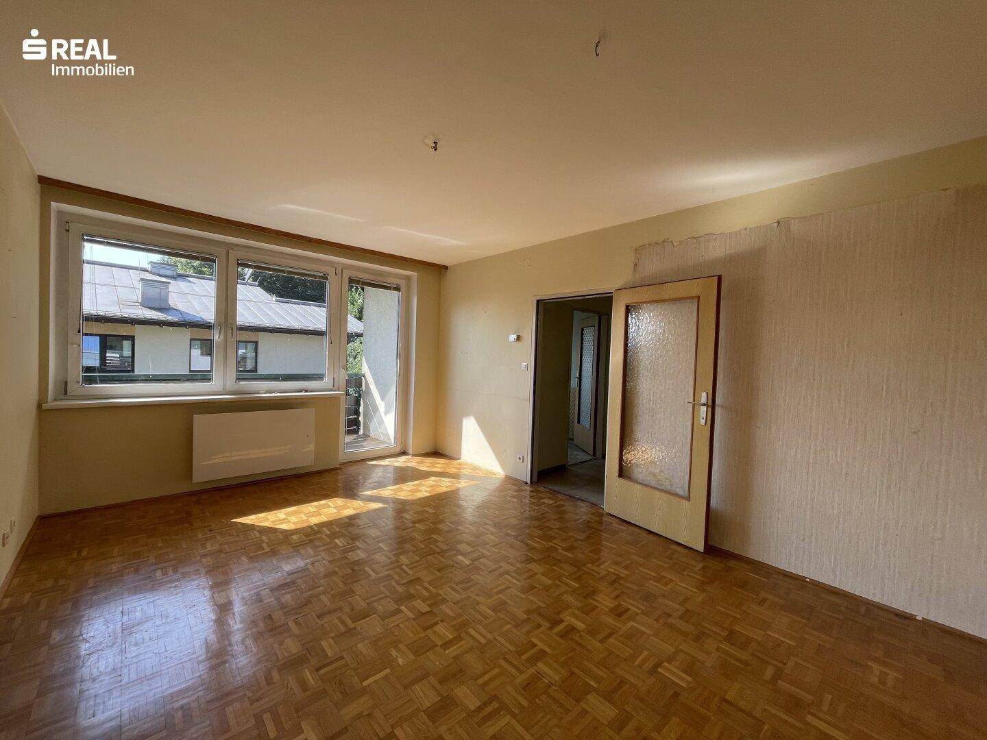 Helles Wohnzimmer mit Parkettboden, großem Fenster und Zugang zu einem Balkon.