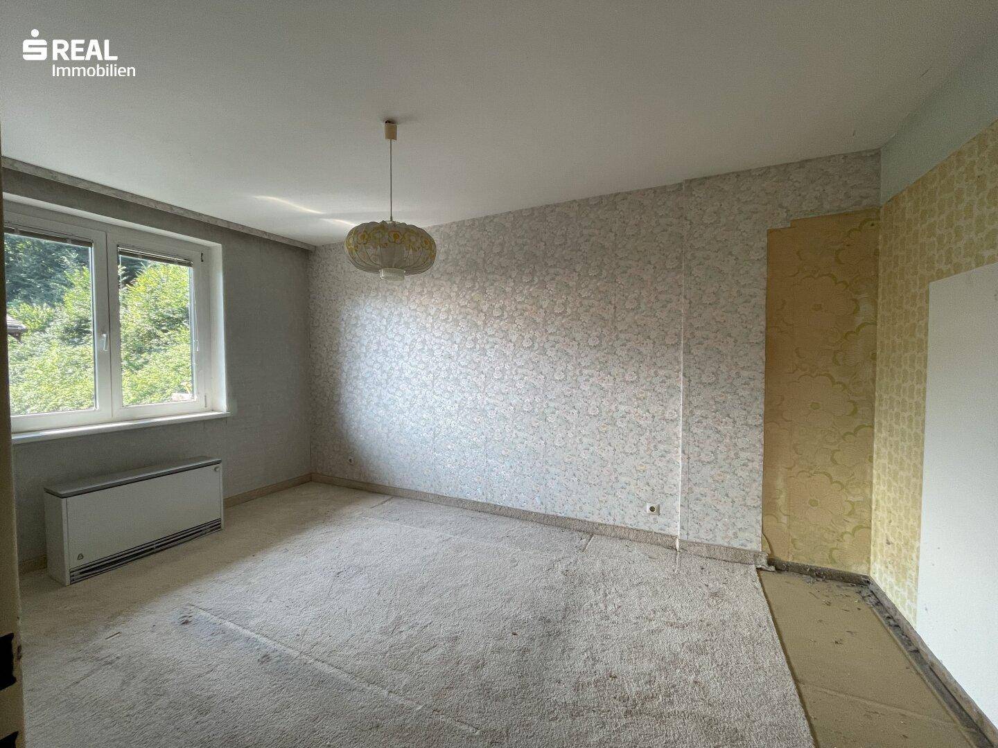 Schlafzimmer mit Fenster und älterer Tapete, das auf eine Neugestaltung wartet.