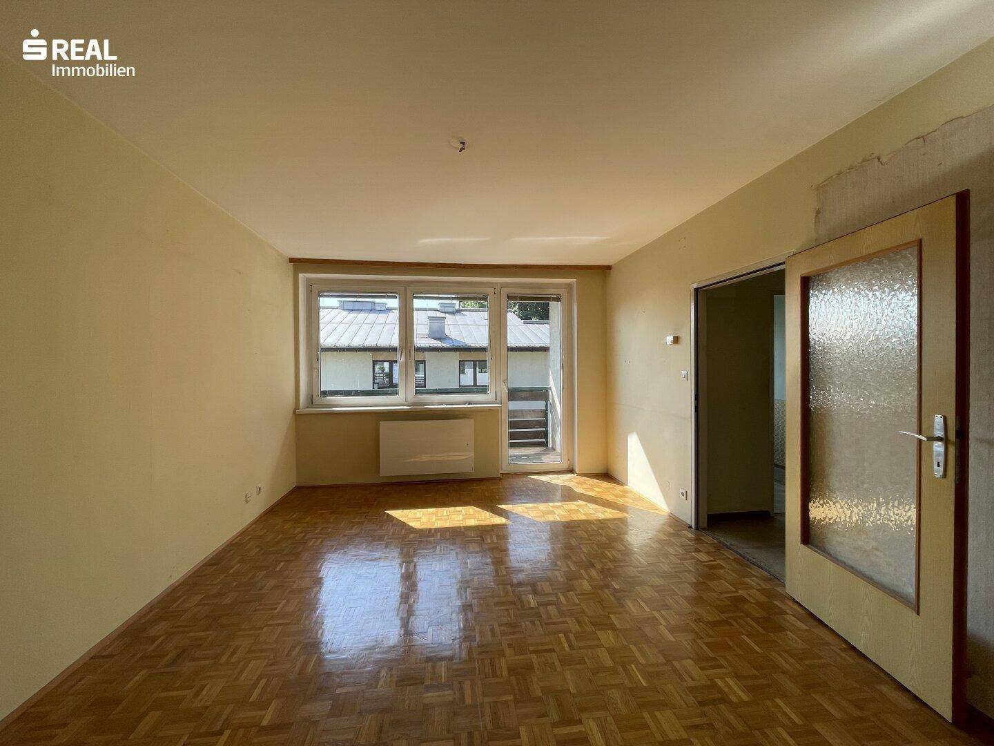 Geräumiges Wohnzimmer mit klassischem Parkettboden und viel Tageslicht durch große Fenster.