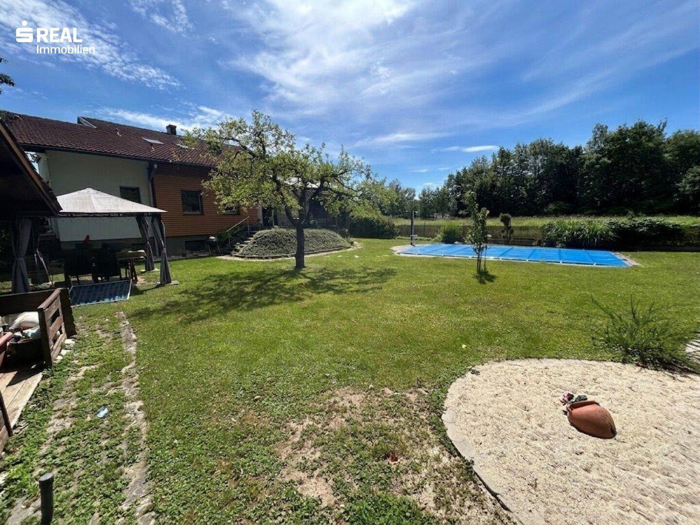 Großer Garten mit Rasenfläche, Sandkasten und Pool, umgeben von Bäumen und dem Wohnhaus im Hintergrund.