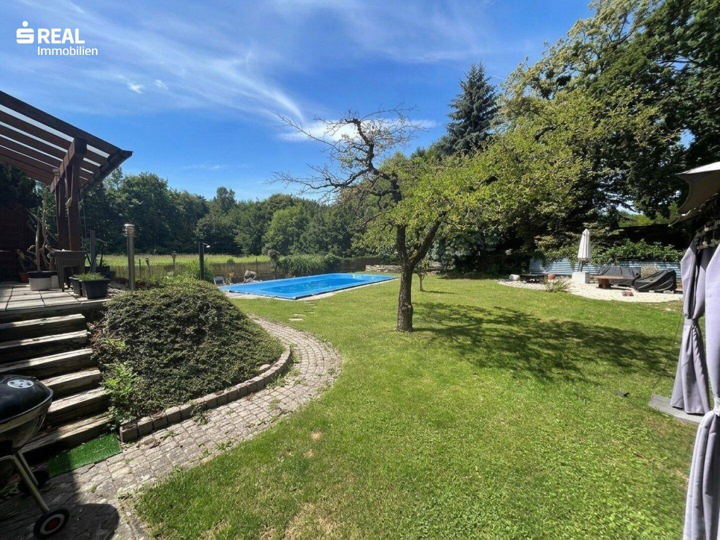 Weitläufiger Garten mit großem Rasenbereich, Schwimmbad und altem Baumbestand unter blauem Himmel.