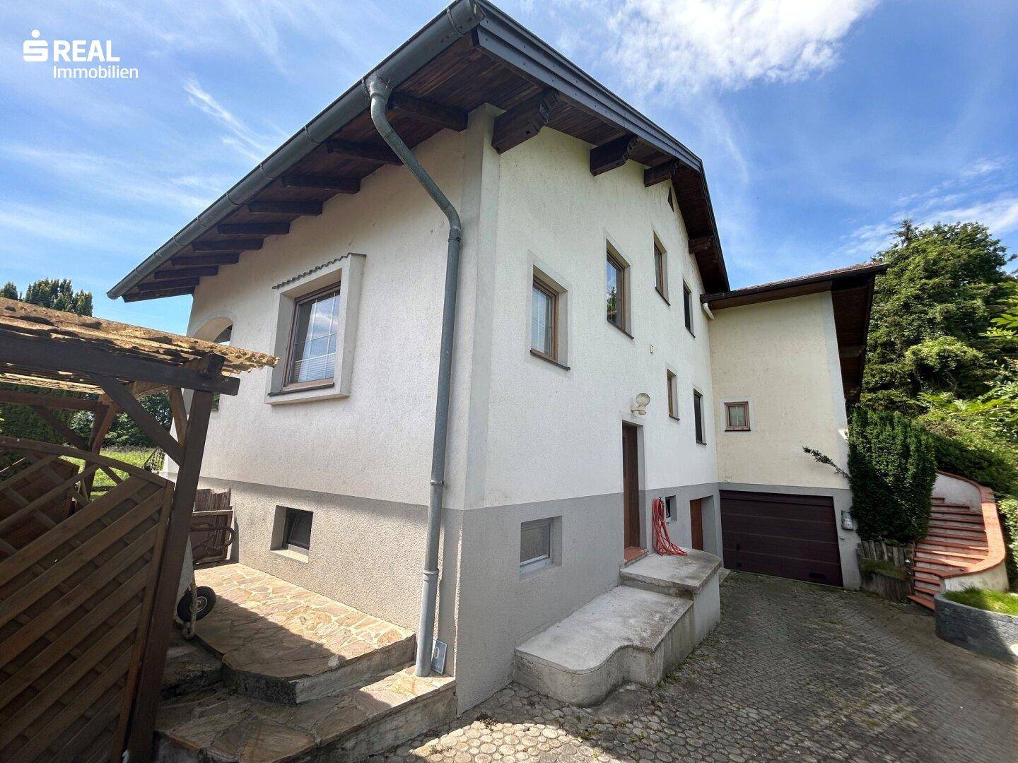 Seitenansicht des Hauses mit weißer Fassade, braunem Garagentor und gepflasterter Auffahrt.