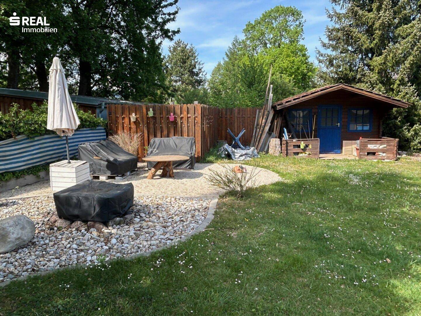 Gemütlicher Sitzbereich im Garten mit Kiesfläche, Gartenmöbeln und einem kleinen Holzschuppen.