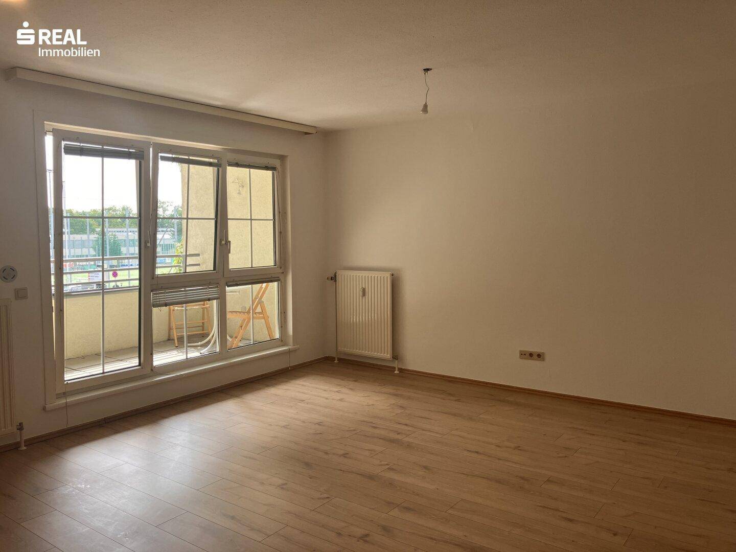 Geräumiges, leeres Wohnzimmer mit Laminatboden und großen Fenstern, die viel Tageslicht hereinlassen.
