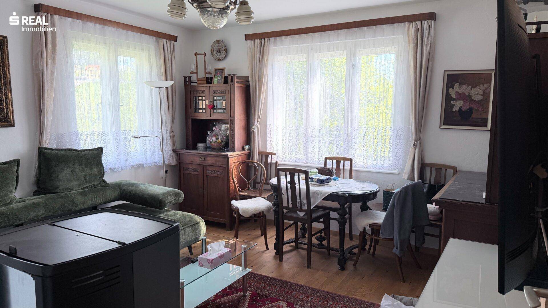 Traditioneller Wohn-Essbereich mit grünem Sofa, Esstisch und klassischen Holzmöbeln, hell durch große Fenster.