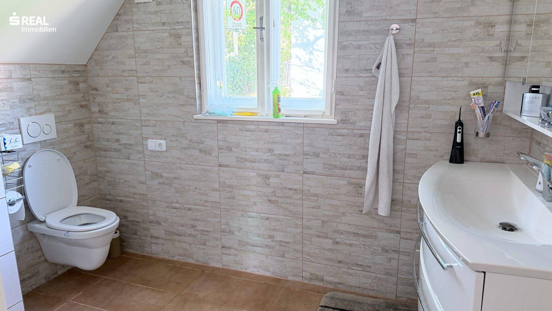 Gefliestes Badezimmer mit Toilette, Waschbecken und Fenster unter einer Dachschräge.