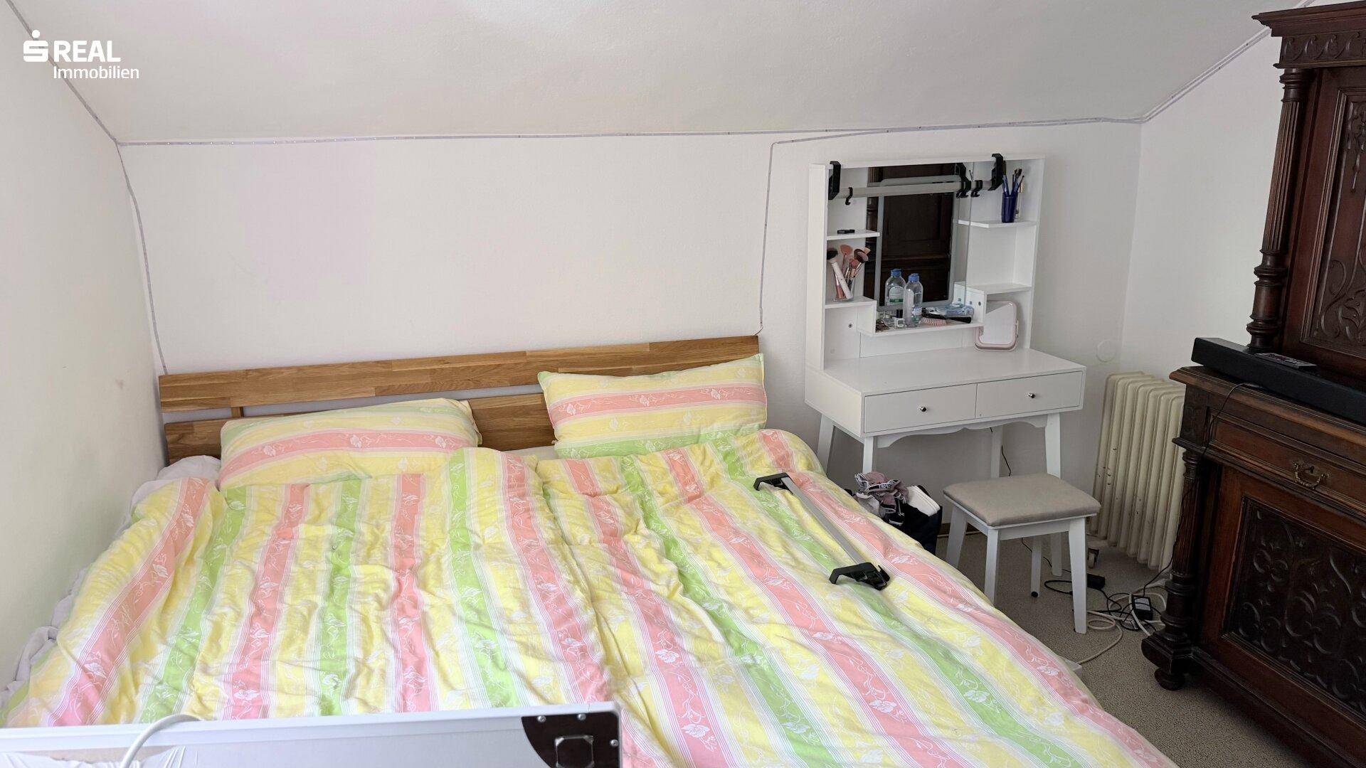 Schlafzimmer mit Bett, Schminktisch und traditionellem Kleiderschrank unter einer Dachschräge.