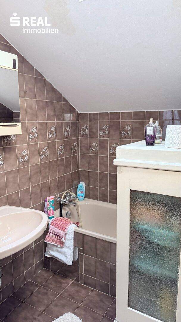 Badezimmer mit braunen Fliesen, Badewanne und Waschbecken unter einer Dachschräge.