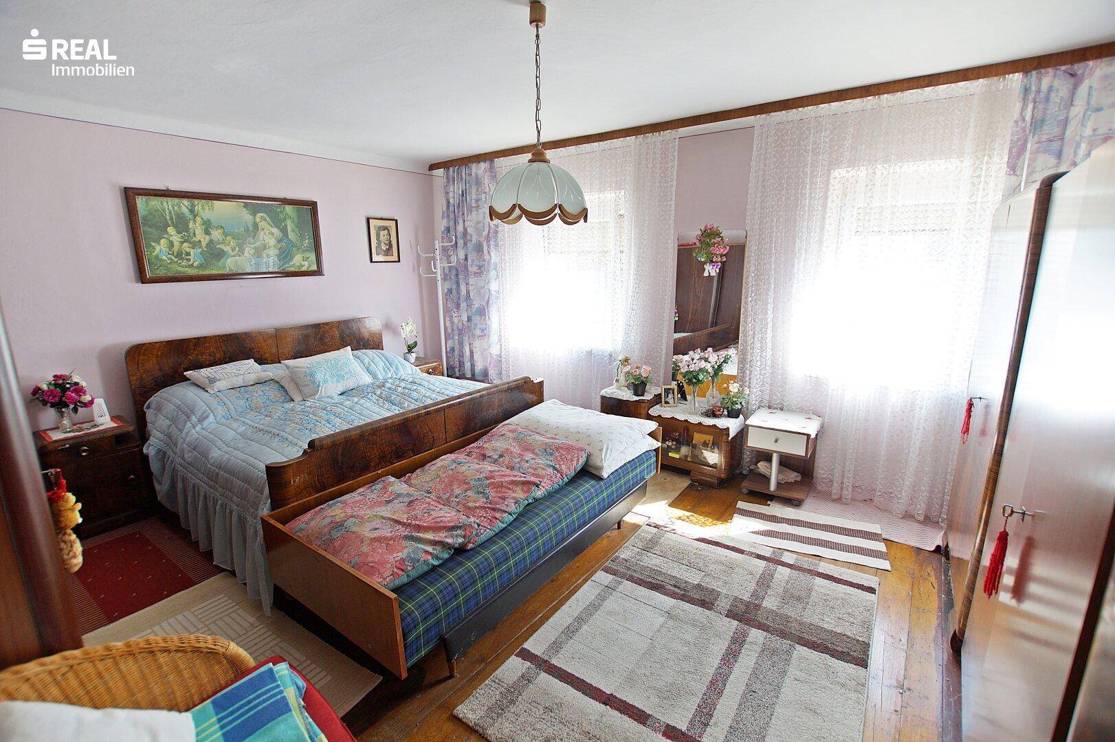 Helles Schlafzimmer mit Doppelbett, Tagesbett und Fenstern mit Vorhängen.