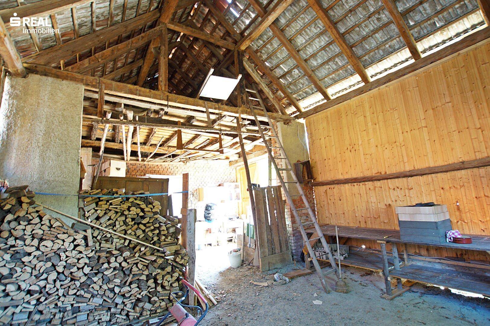 Innenansicht einer Scheune mit Holzbalken, Holzstapel und Leiter.