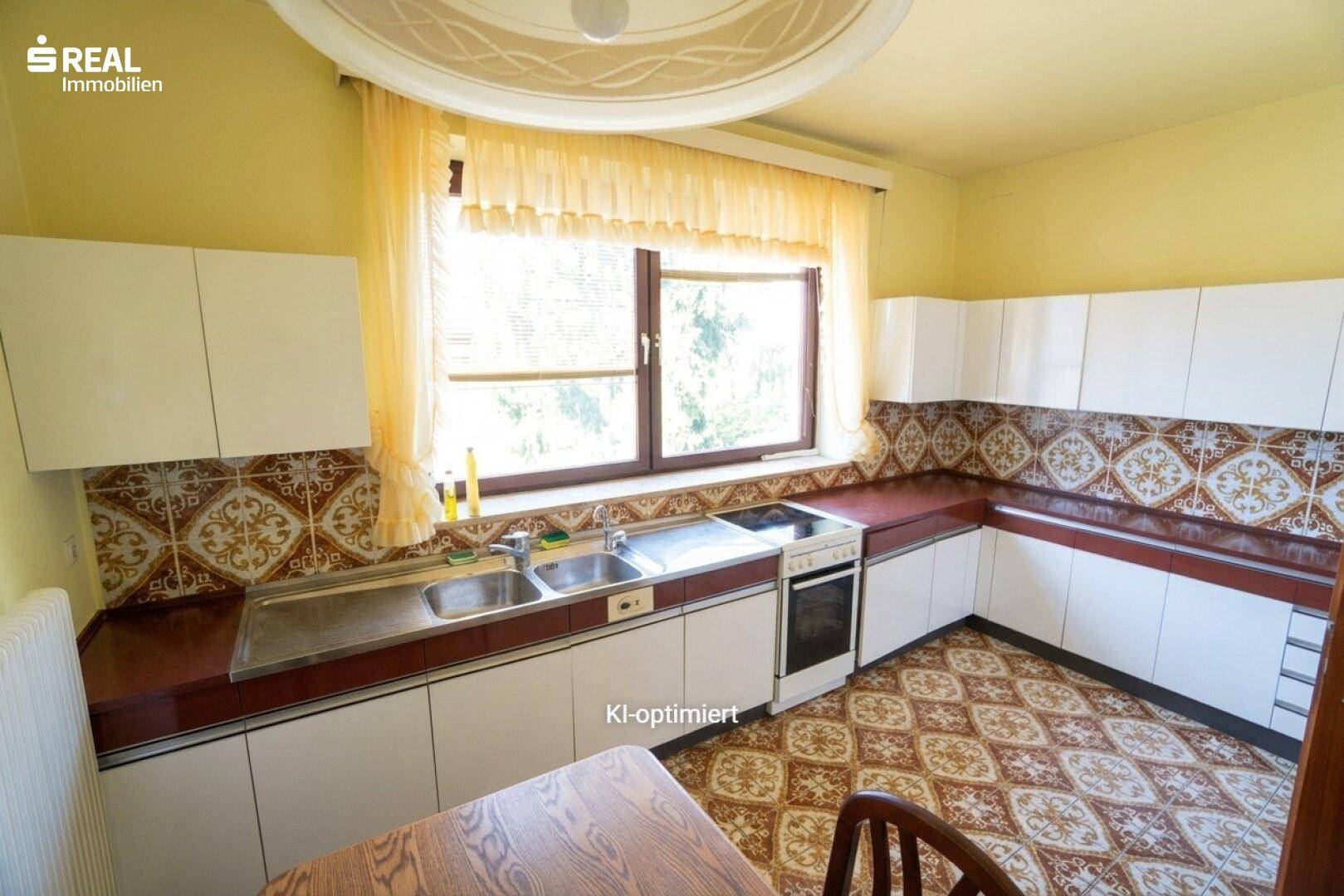 Funktionale Küche mit weißen Schränken, gemustertem Fliesenboden und Essbereich am Fenster.