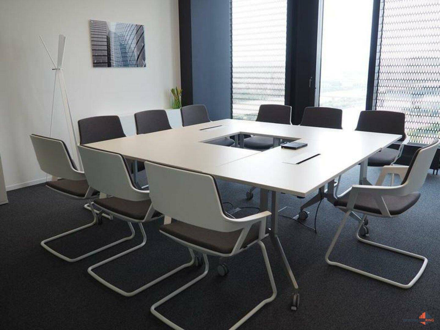 Heller Konferenzraum mit großem quadratischem Tisch und modernen Stühlen, ideal für Besprechungen.