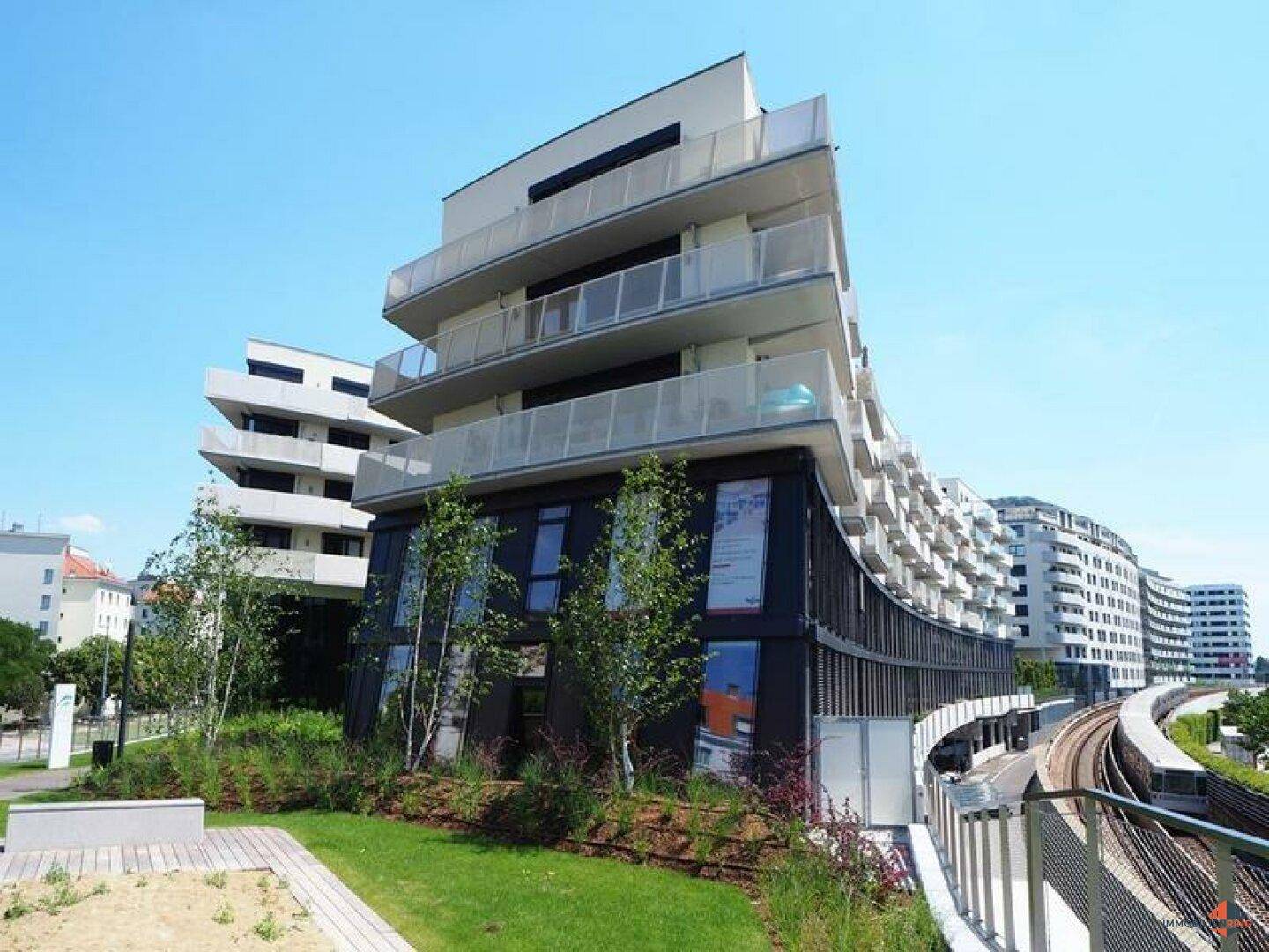 Modernes Mehrfamilienhaus mit geschwungener Fassade, zahlreichen Balkonen und angrenzenden Bahngleisen.