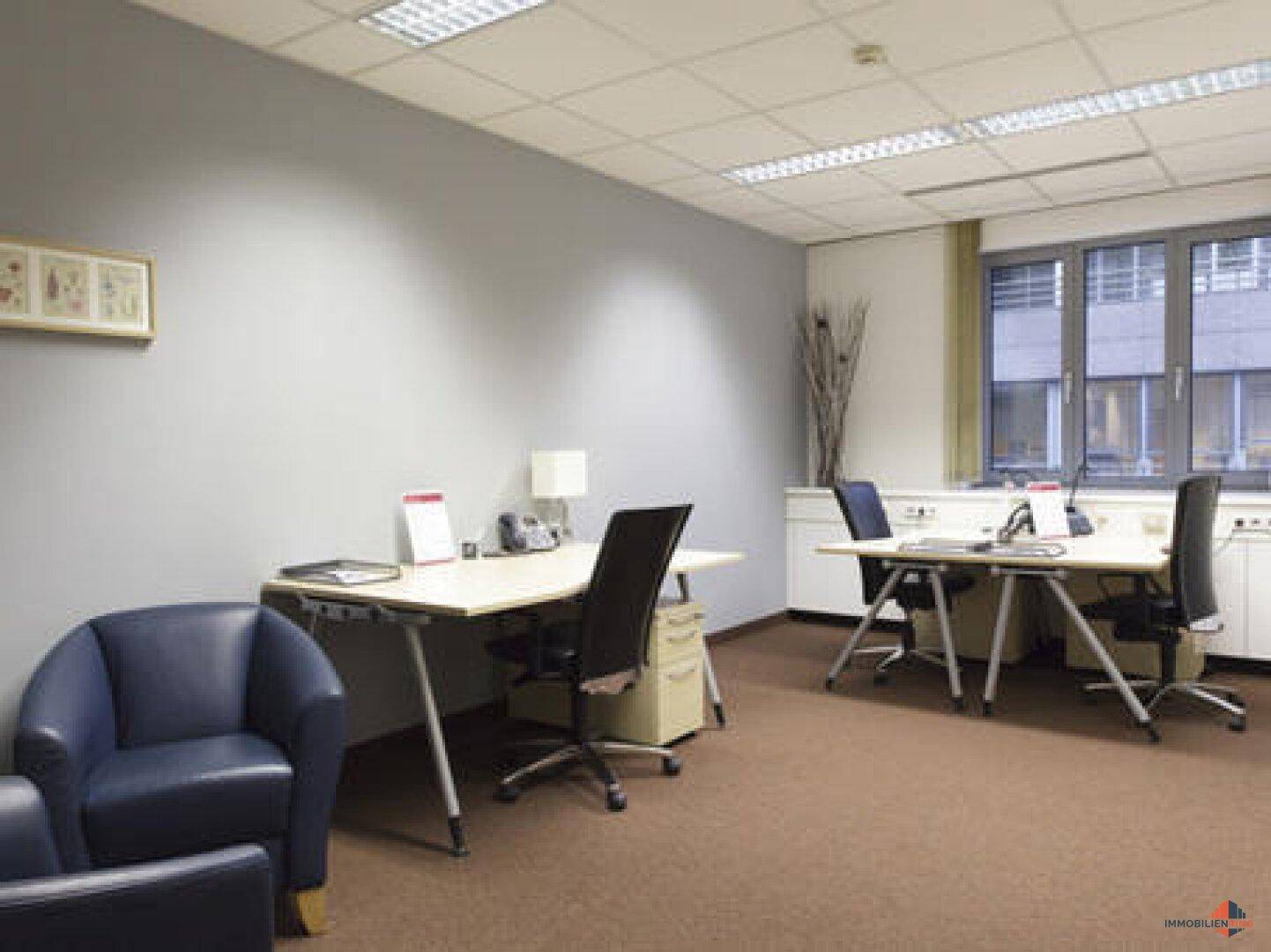 Geräumiges Büro mit mehreren Arbeitsplätzen, Teppichboden und einem großen Fenster.