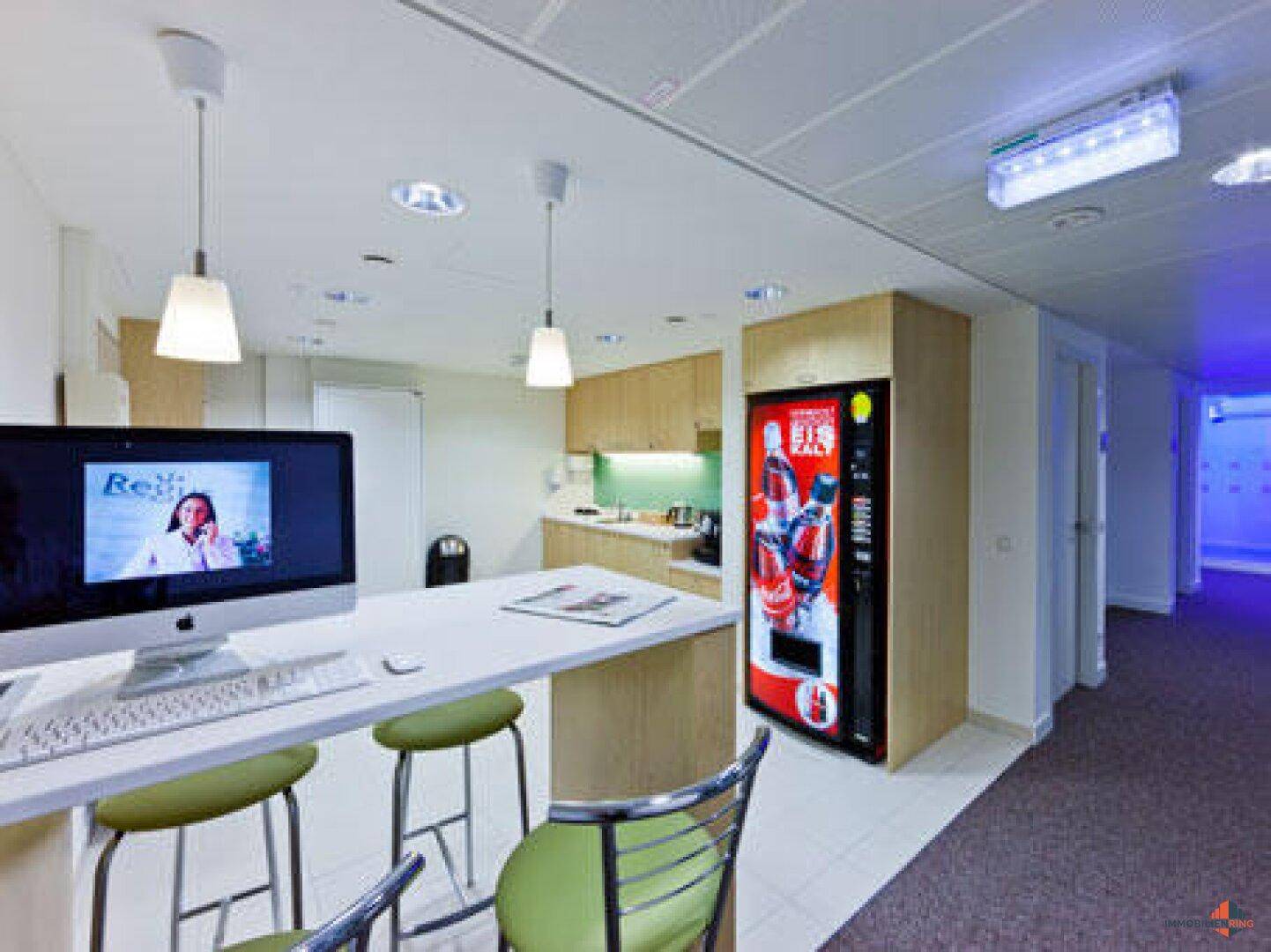 Offener Bürobereich mit Arbeitsplatz, integrierter Küchenzeile, Verkaufsautomat und hohen Decken.