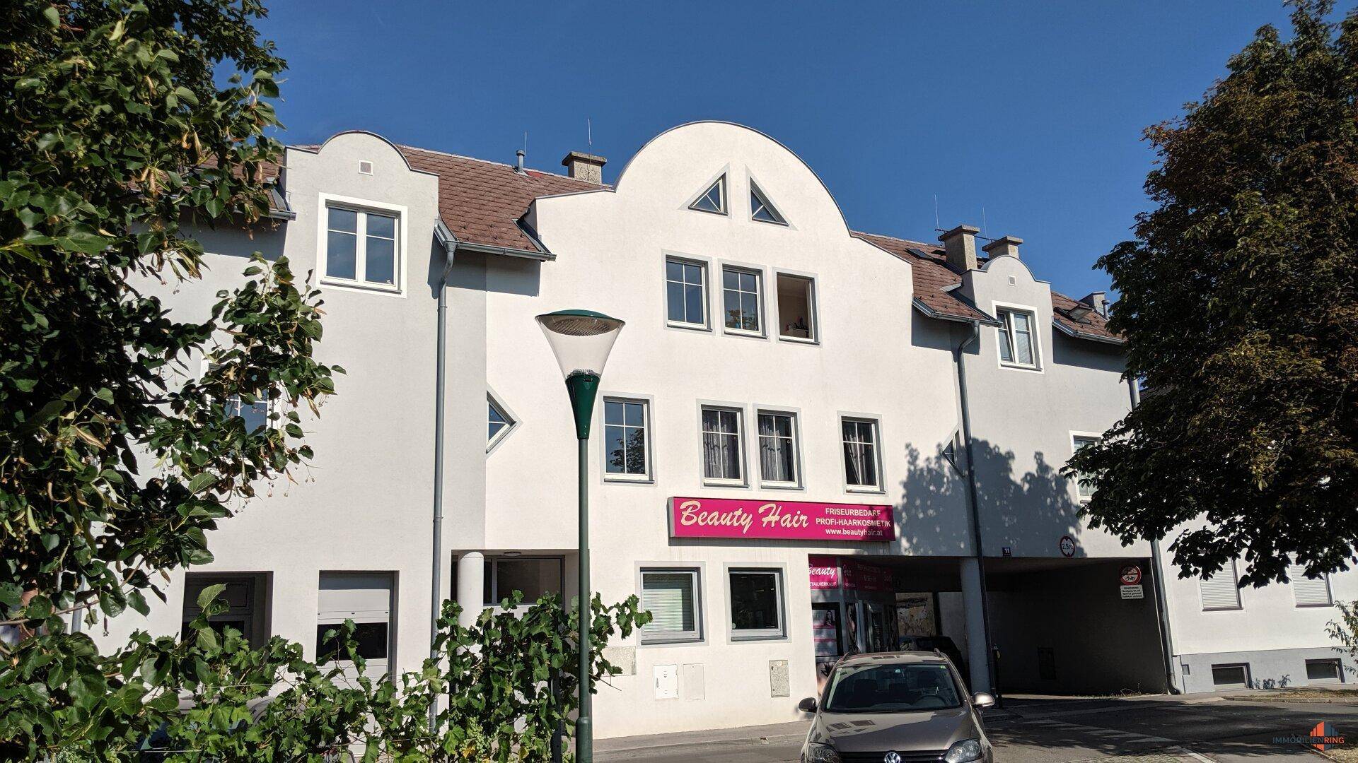 Fassade eines Mehrfamilienhauses mit Geschäftsräumen im Erdgeschoss und Bäumen, gepflegtes Erscheinungsbild.