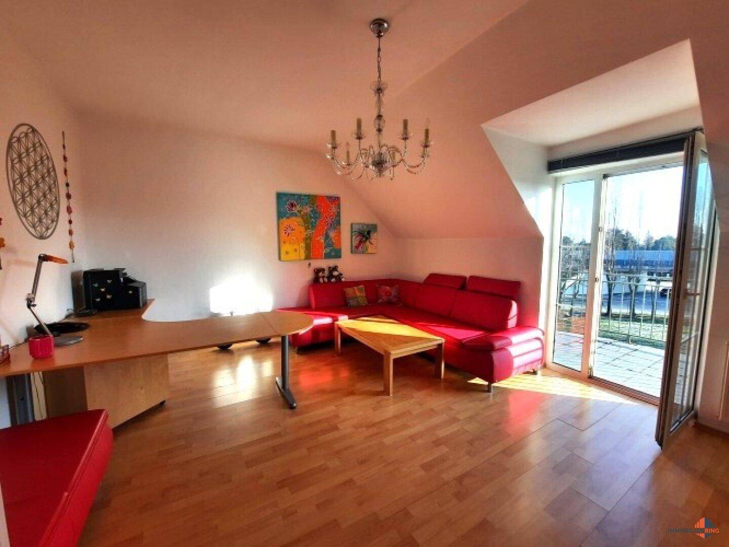 Helles Wohnzimmer im Dachgeschoss mit rotem Sofa, Holzboden und Zugang zu einem Balkon.