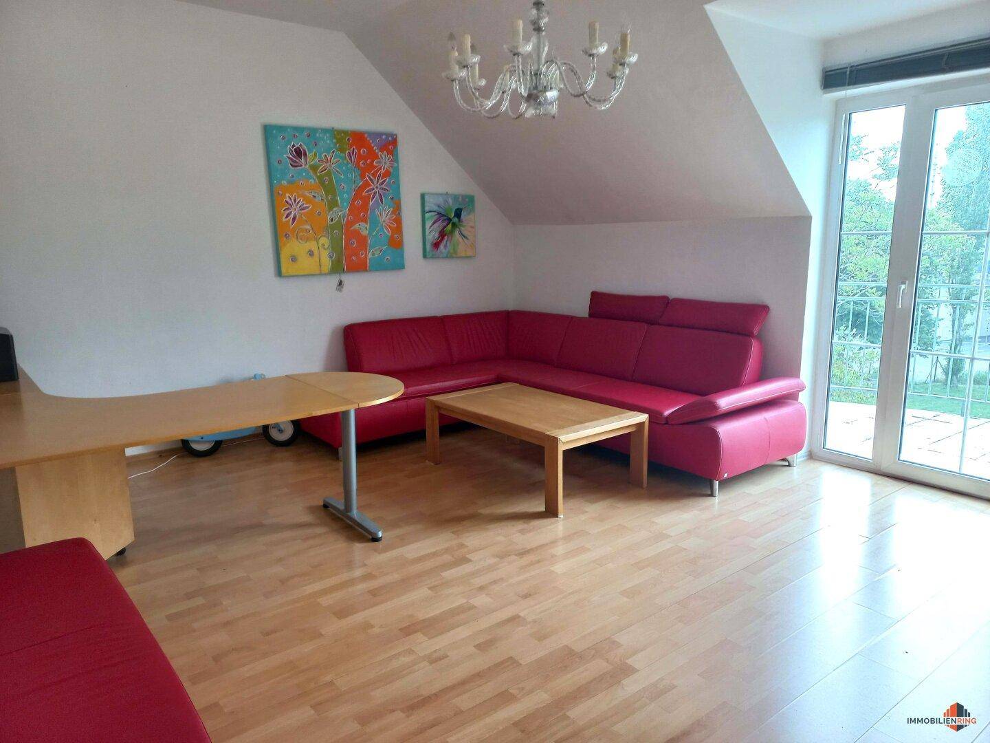 Gemütliches Wohnzimmer im Dachgeschoss mit rotem Ecksofa, Holzboden und Blick ins Grüne.