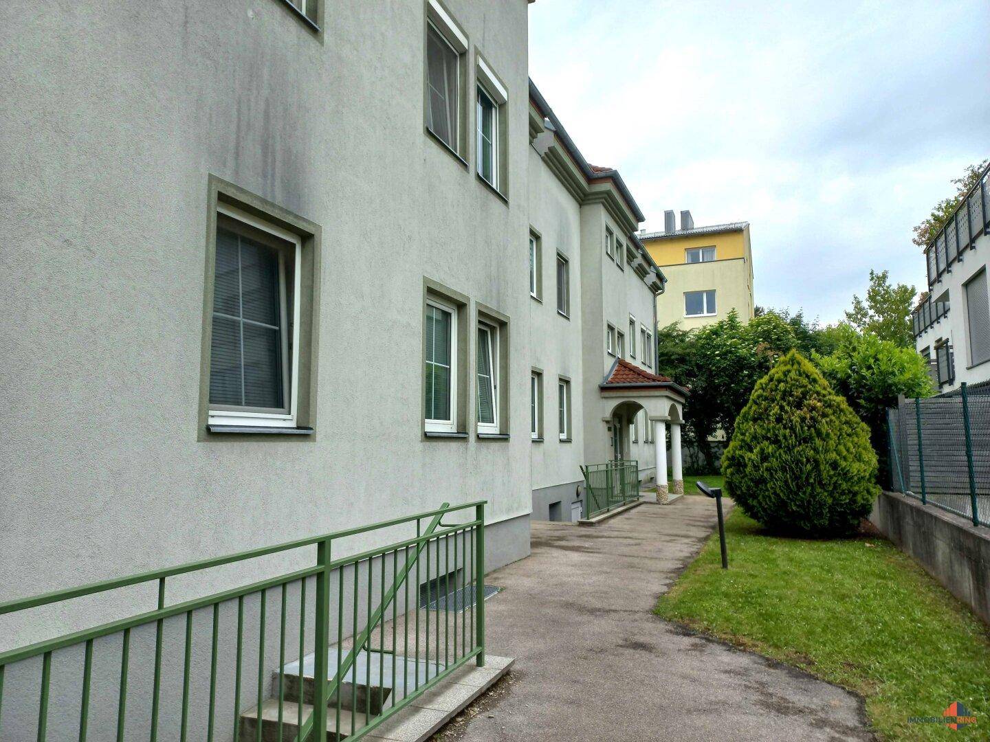 Graue Fassade eines Mehrfamilienhauses mit grünem Geländer und Zugang zum Eingangsbereich.