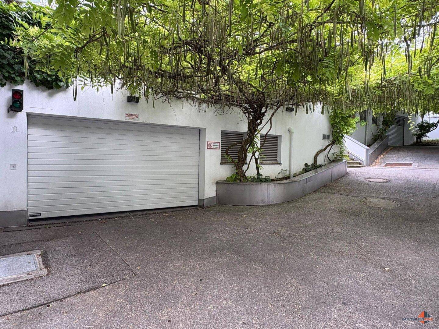 Garageneinfahrt mit automatischem Tor, umgeben von üppiger Vegetation.