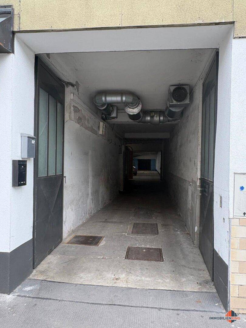 Langer, schmaler Zugangsbereich mit Betonboden und sichtbaren Lüftungsrohren an der Decke.