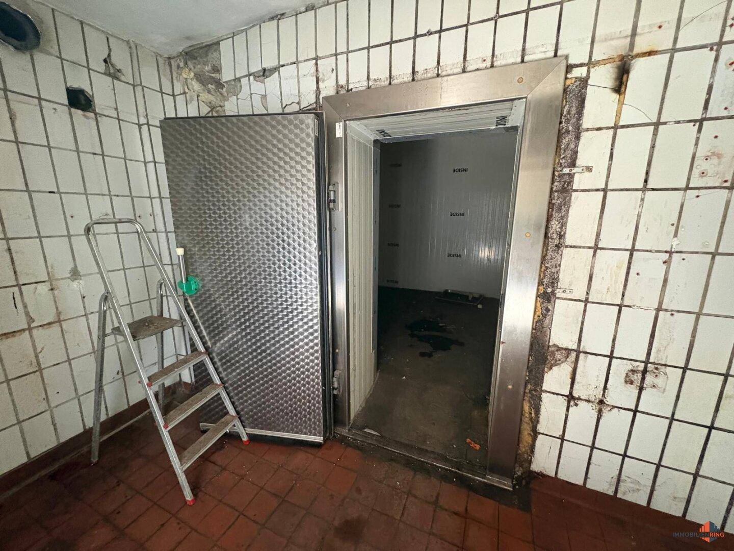 Kühlraum mit Edelstahl-Tür, weißen Wandfliesen und Terrakotta-Boden, bereit zur Nutzung.