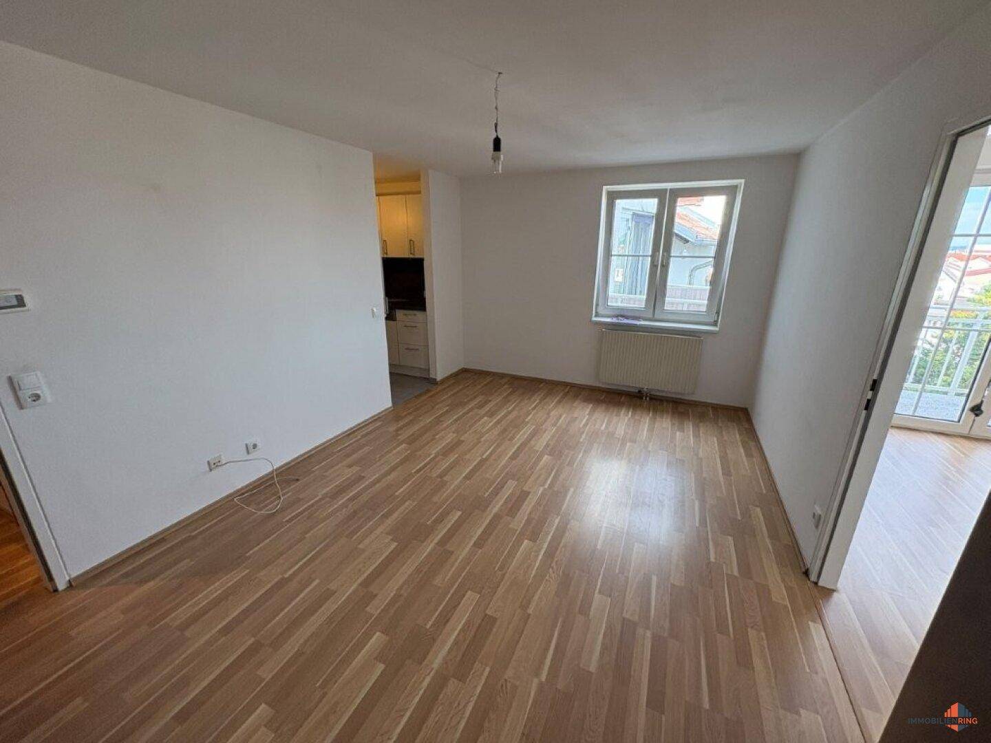 Wohnbereich mit Parkettboden und Fenster, offen zu einer kleinen Küchenzeile, hell und einladend.