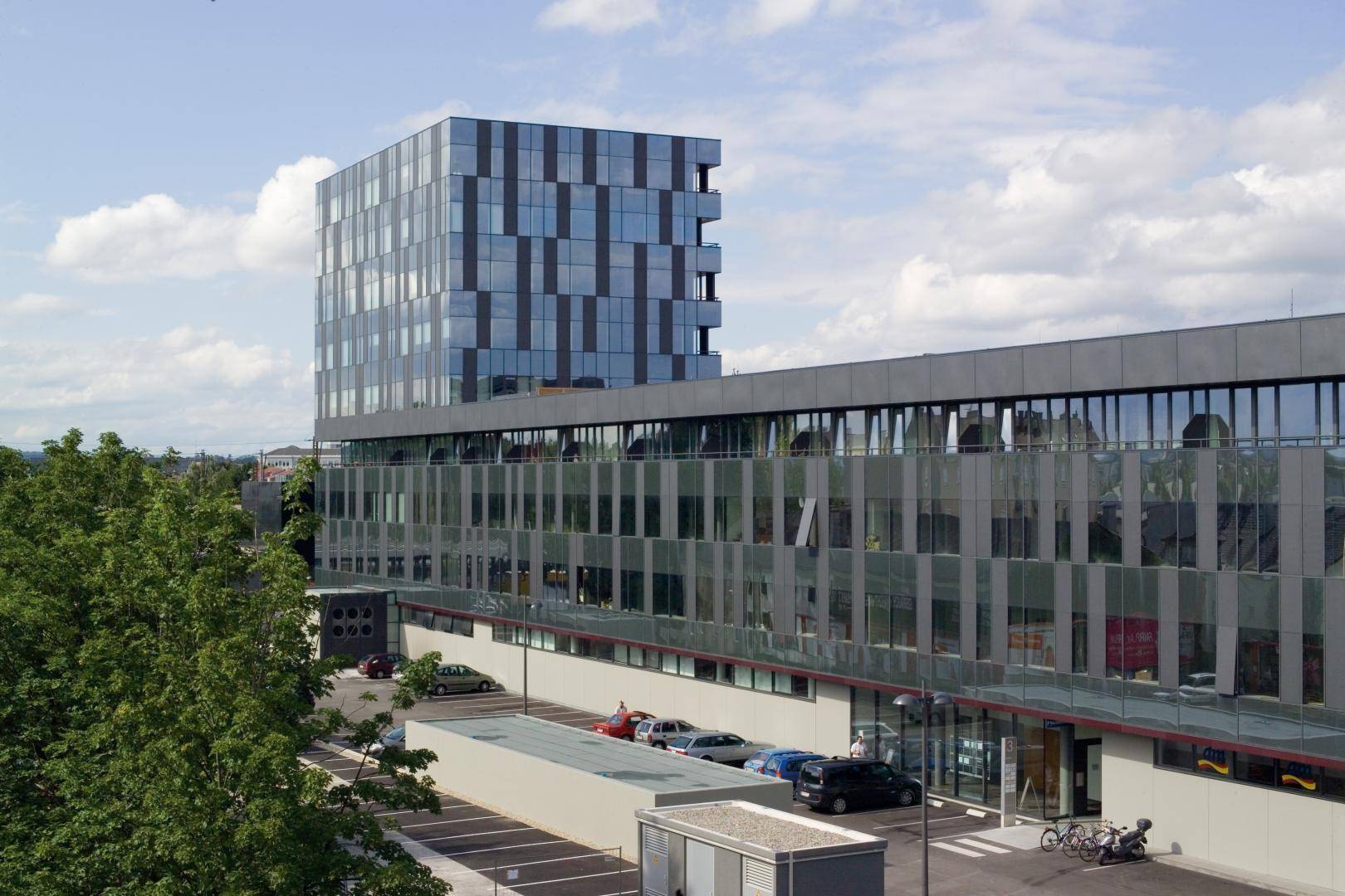 Moderne Bürogebäude mit markanter Glasfassade und umliegenden Parkplätzen unter einem bewölkten Himmel.