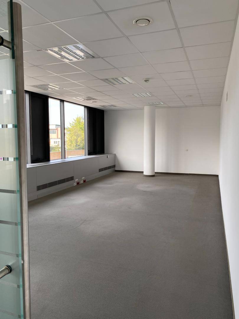 Blick durch eine Glastür in einen hellen Bürobereich mit Vinylboden, großen Fenstern und einer Säule.