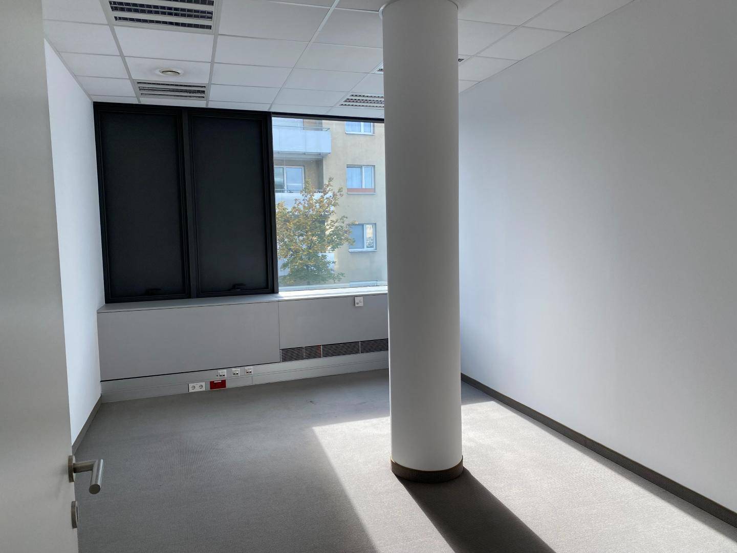 Helles Büro mit einer zentralen Säule, großen Fenstern und Teppichboden, ideal für Einzelbüros.