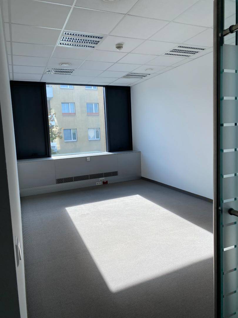 Kleineres, helles Büro mit Teppichboden und Blick auf ein gegenüberliegendes Gebäude, durch eine Glastür zugänglich.