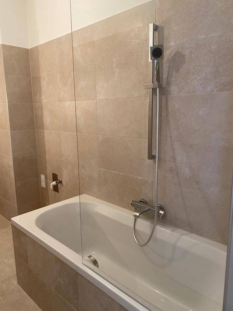 Helles Badezimmer mit Badewanne und Duscharmatur, umgeben von beigefarbenen Fliesen.