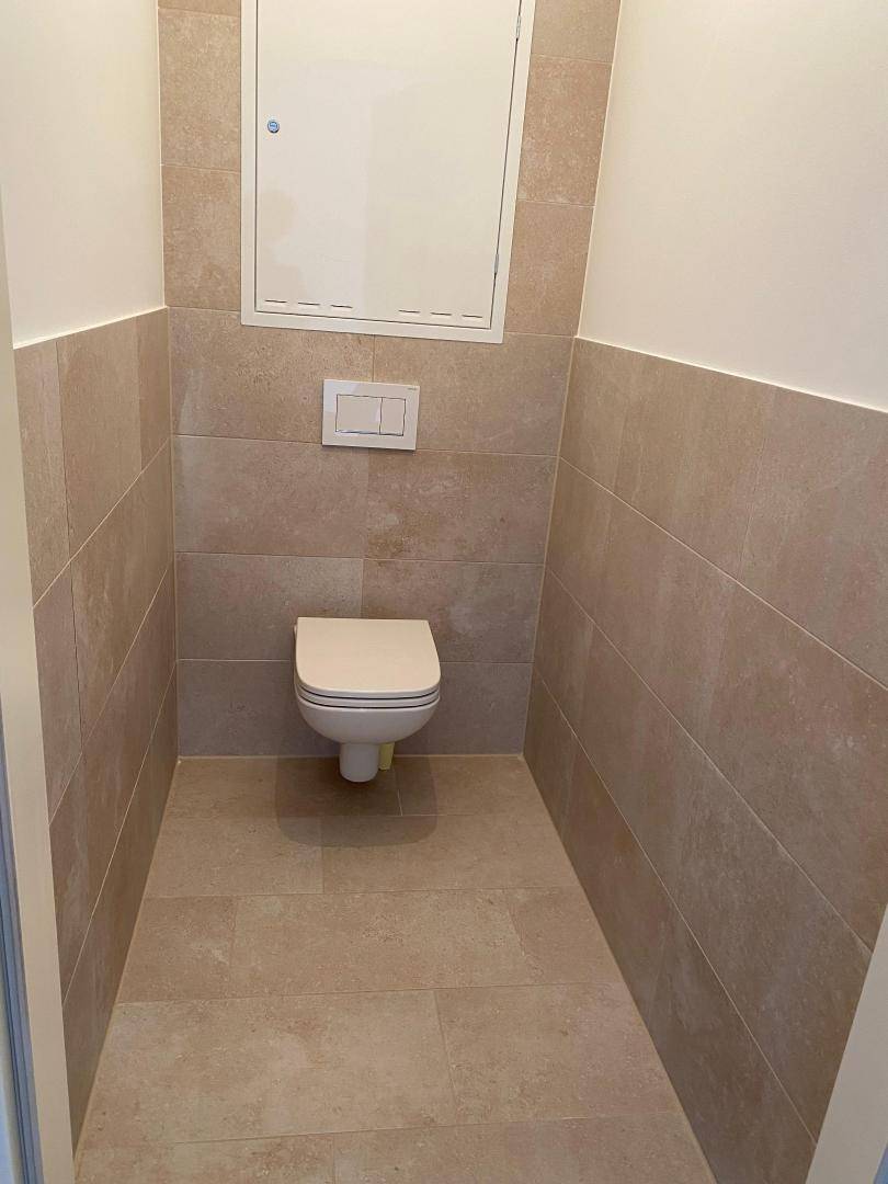 Separates WC mit wandhängender Toilette und beigefarbenen Fliesen an Wänden und Boden.