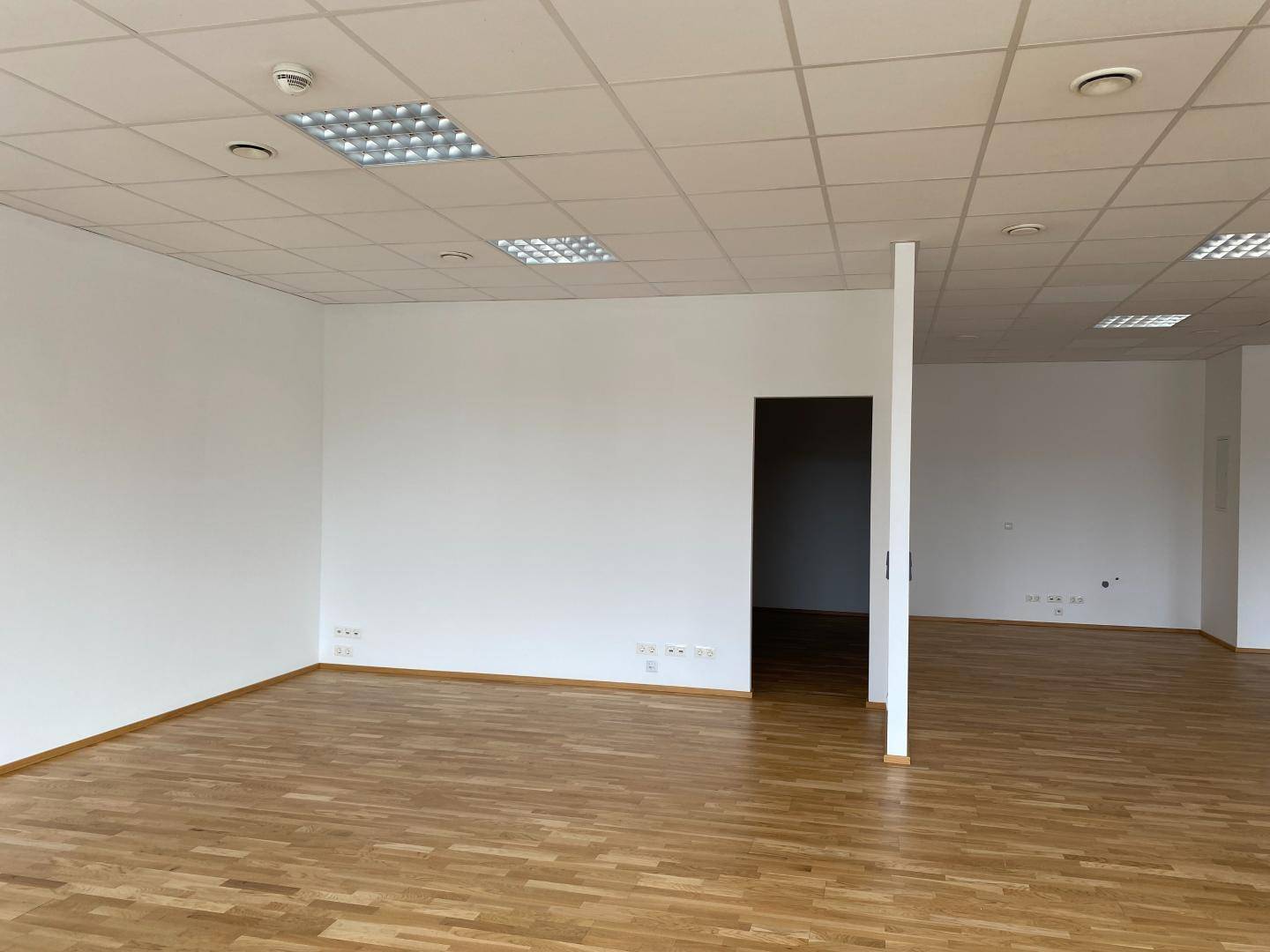 Offener Bürobereich mit Holzboden, weißen Wänden und einem Durchgang zu einem weiteren Raum.