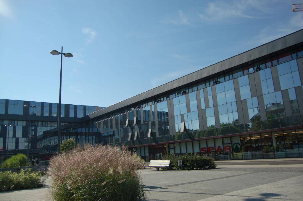 Moderne Gebäudefassade mit großen Glasflächen und einem Supermarkt im Erdgeschoss, umgeben von Grünflächen.