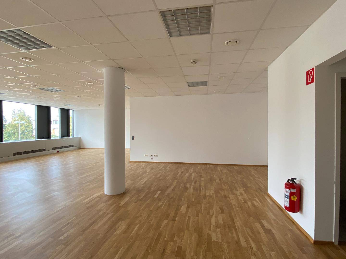 Leerer Bürobereich mit Holzboden, weißen Wänden und einem Feuerlöscher an der Wand.