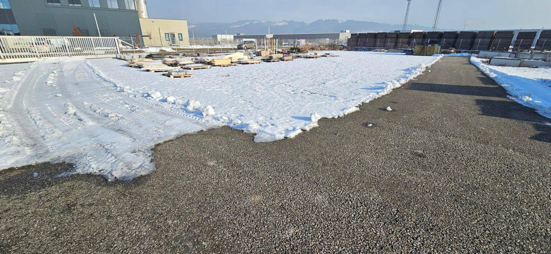 Große Freifläche mit Schotterboden, teilweise schneebedeckt, im Hintergrund sind Berge sichtbar.