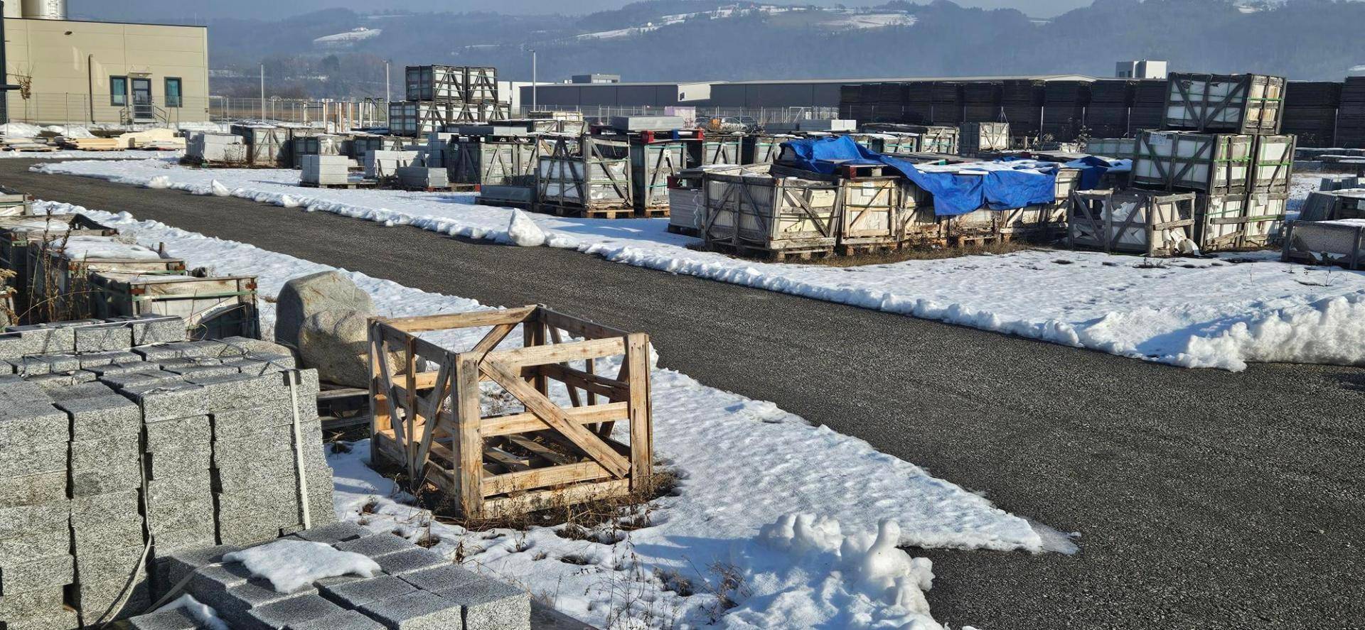 Industrielle Außenanlage mit gelagerten Steinplatten, Holzkisten und einem Gebäude im Hintergrund, teilweise schneebedeckt.