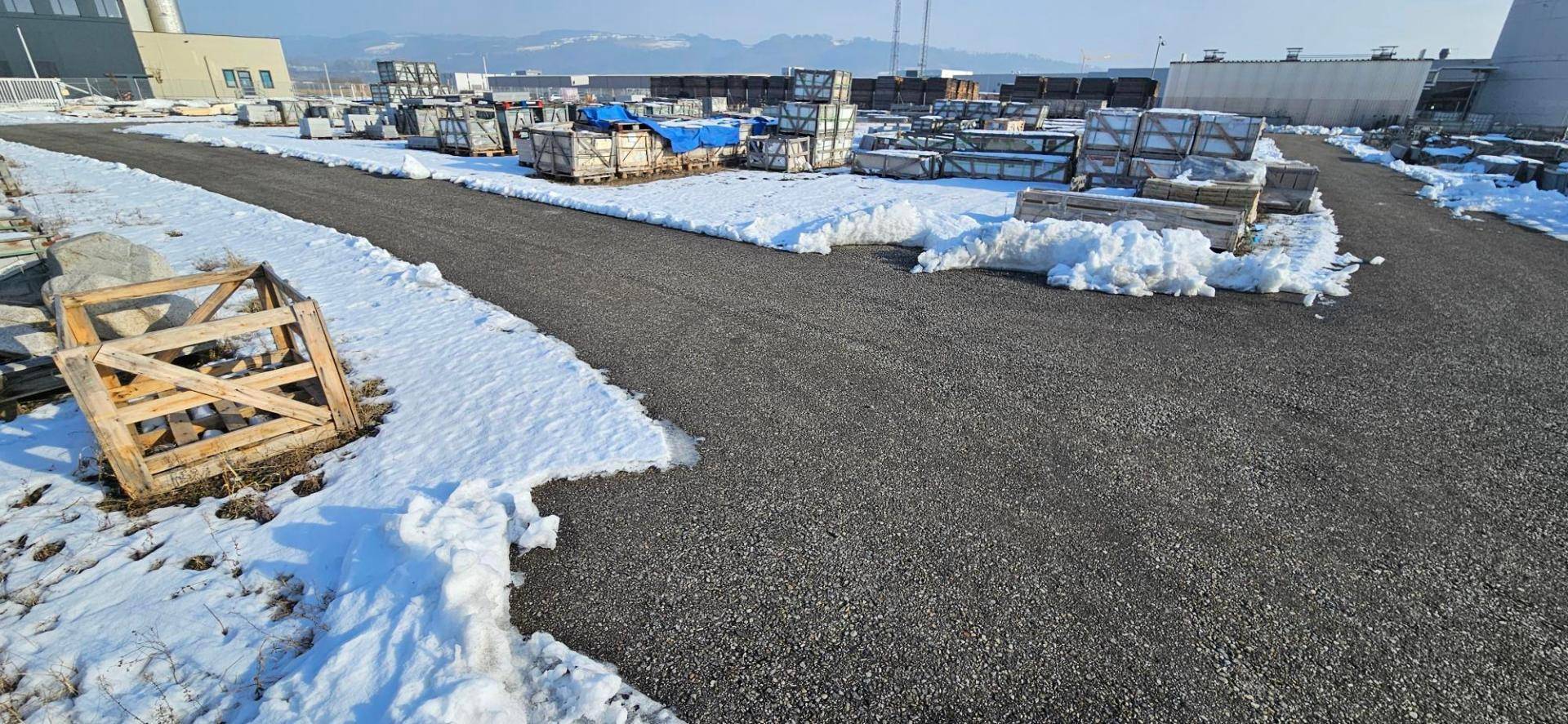 Weitläufige Außenfläche mit Schotterweg, Schneeresten und gelagerten Materialien in Holzkisten.