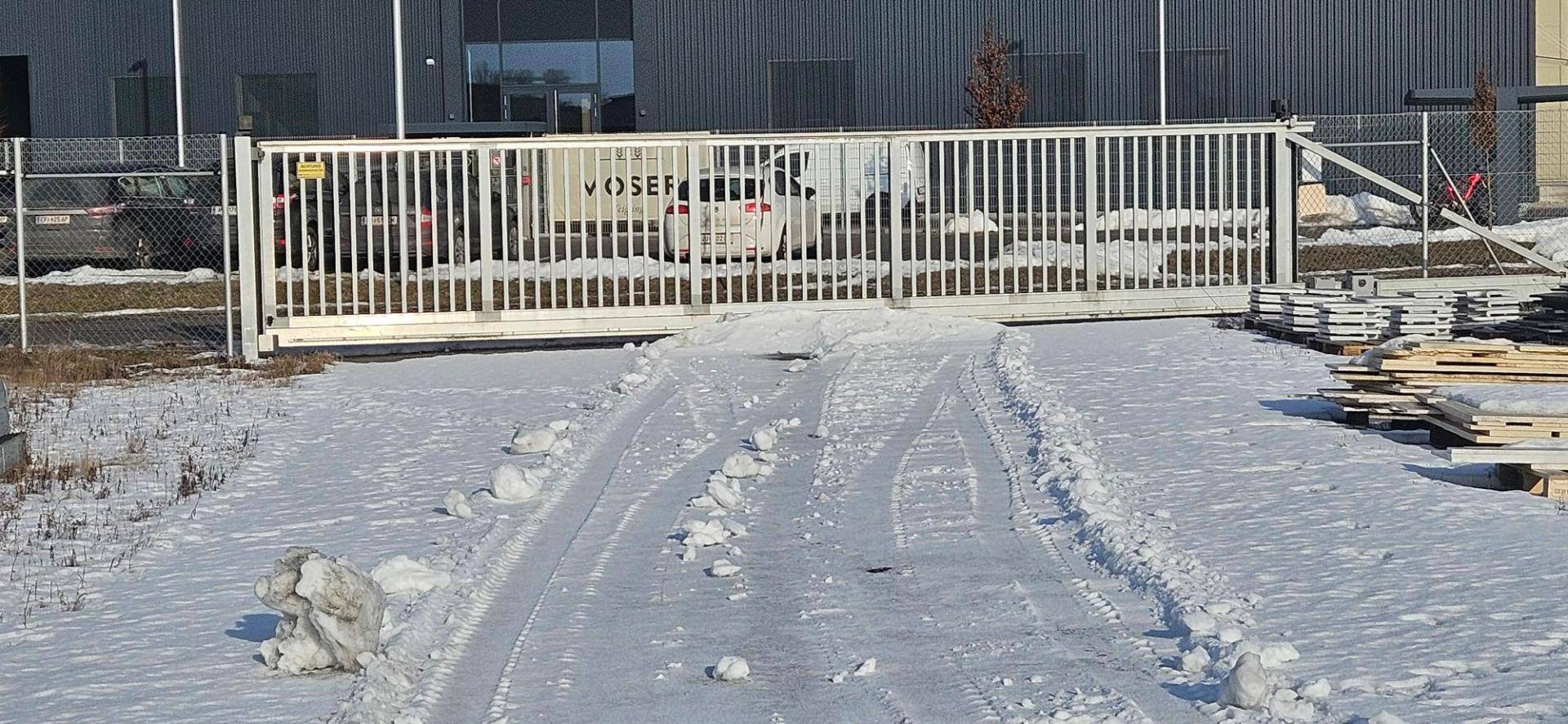 Großes weißes Schiebetor auf einem schneebedeckten Zufahrtsweg zu einem Industriegelände.